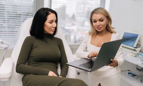 Aesthetic clinic consultation where a practitioner in a white outfit shows treatment options on a laptop to a client seated on a treatment chair in a modern medspa setting.