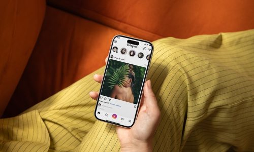 Hand holding iPhone displaying an Instagram feed against an orange background.