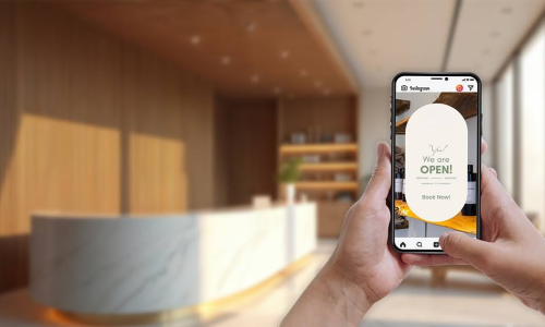 Person holding a smartphone displaying an Instagram Story with a "We are OPEN! Book Now!" message, set against a blurred modern reception or salon interior